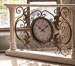 The photo depicts an elegant mantel clock with a vintage appearance. The clock features a classic face against a white background. The clock face is surrounded by an elaborately decorated frame with swirling, baroque motifs. The frame has a weathered, white color that adds to its antique character. The clock is placed on what appears to be a windowsill, with sunlight streaming through a window with a weathered frame. This gives the scene a warm, inviting atmosphere and accentuates the texture of the clock's metal frame.