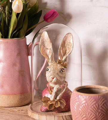 Een figuur van een konijn, gekleed in een outfit compleet met een vest en een strikje. Het konijnenbeeldje is ingekapseld in een transparante glazen stolp die bovenop een houten basis staat. Aan de linkerkant van de konijnenweergave staat een roze vaas met een arrangement van tulpen, wat de scène een vleugje lentefrisheid geeft. Direct naast de stolp staat een keramische mok, die de roze tinten van de vaas weerspiegelt en voorzien is van een tribalachtig patroon, wat bijdraagt aan de warme, rustieke esthetiek van de scène.