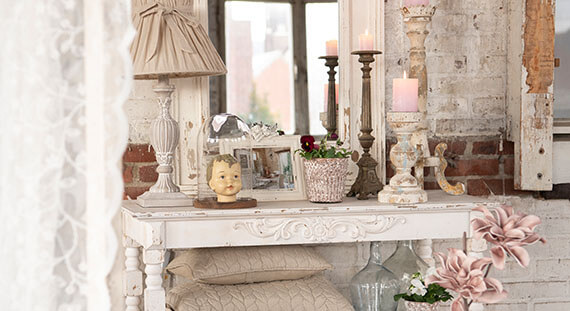 Ein stimmungsvoll eingerichteter Innenraum im Vintage- oder Shabby-Chic-Stil. In der Mitte steht ein weißer Holztisch, verziert mit eleganten Holzschnitzereien. Auf dem Tisch sind verschiedene dekorative Gegenstände zu sehen: Kerzen auf hohen Kerzenständern, eine Glasvase mit Blumen, eine kleine Engelsfigur und verschiedene andere Trödel, die ein antikes Aussehen haben. Eine kleine spiegelnde Oberfläche reflektiert einen Teil der Szene. Hinter dem Tisch ist eine Wand mit teilweise freigelegten Ziegeln sichtbar, was zum rustikalen Charme beiträgt. Es hängt auch eine Tischlampe mit einem klassischen Lampenschirm. Links ist ein Stück eines weißen Vorhangs zu sehen und rechts im Vordergrund befinden sich Blumen im gleichen Farbschema. Die gesamte Kulisse strahlt eine warme, gemütliche Atmosphäre aus und scheint im natürlichen Tageslicht aufgenommen zu sein.