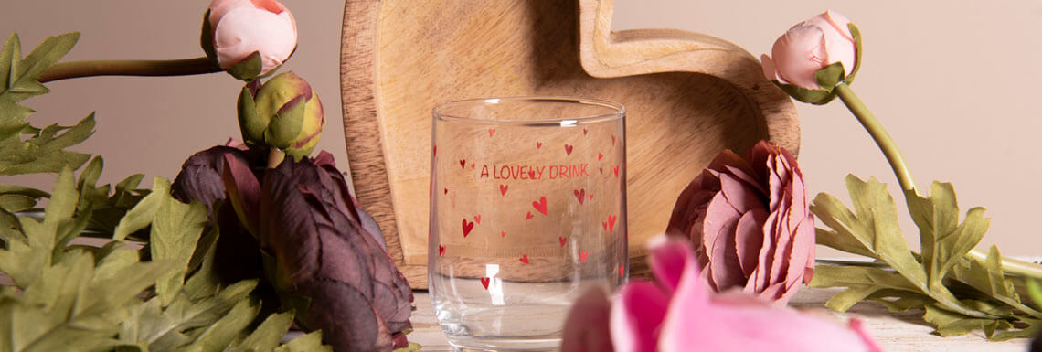 A close-up of an atmospheric table setting with a romantic theme. At the center of the image stands a clear drinking glass etched with the words 'A LOVELY DRINK' surrounded by small red hearts. Behind the glass, a portion of a wooden cutting board is visible, featuring a heart shape cut out at the top. Artificial flowers are also laid on the table, including pink peonies and green leaves that provide a soft and elegant contrast against the neutral background. The color palette is gentle, with pink hues of the flowers nicely complementing the natural wood tones and the neutral glassware. The whole suggests a setting for a loving occasion or Valentine's Day theme.