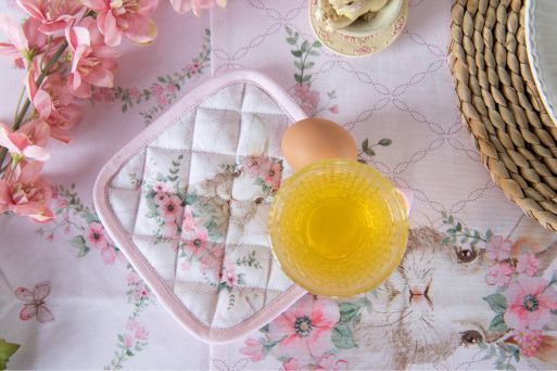 Een pannenlap met schattige, roze bloemen en zoete patronen, uit de textielserie Floral Easter Bunny, ligt naast een bijpassend tafelkleed. Op de tafel liggen ook een glas drinken en bloemen.
