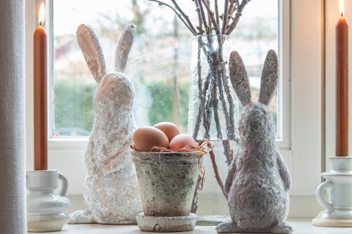 Twee grote paashazen, een witte en een kleinere grijze, kijken uit het raam. In het midden staat een decoratieve emmer gevuld met eieren. Aan beide uiteinden bevinden zich kandelaren met brandende kaarsen, wat zorgt voor een complete paassfeer.