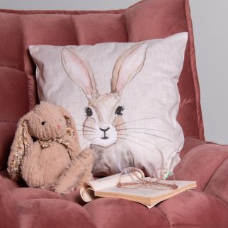 On a powder pink chair lies an Easter cushion with an illustration of an Easter bunny, accompanied by a stuffed animal and a book.