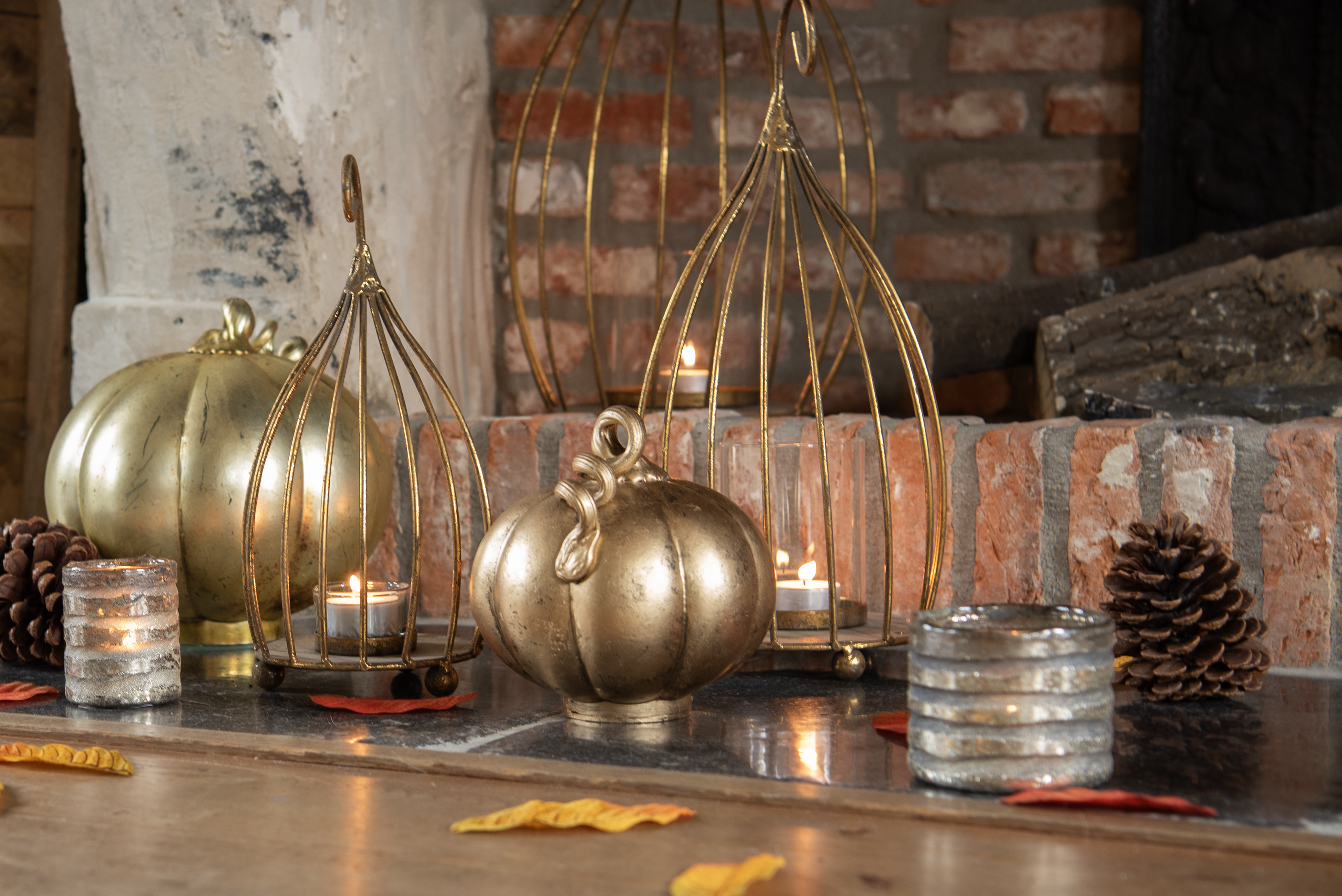 A cozy and warm autumn or Halloween decoration on a wooden table or mantle, against a backdrop of a brick wall with visible traces of peeling plaster. Central to the setting is a large, metal pumpkin with a rich golden hue, along with a smaller, similarly designed pumpkin. Both pumpkins have a polished surface that softly reflects the light from the surrounding candles.

On the left side of the composition is a metal candle holder with an open structure, allowing the light from the burning candles to spread atmospherically throughout the space. Another similar candle holder is visible on the right, and both contribute to the warm glow of the setting.

Scattered across the table are small silver candle holders with burning candles inside, enhancing the overall coziness. Natural elements such as pinecones and autumn leaves with rich red and gold tones have also been added to the decoration, strengthening the association with autumn and adding a sense of natural beauty to the whole setup.