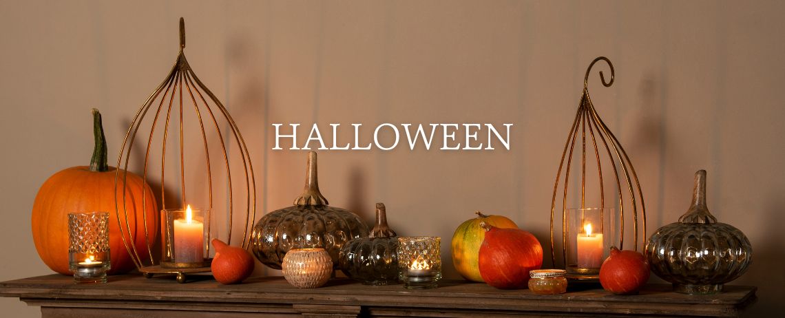 A warm and inviting Halloween decoration set up on a wooden surface. Centered in the image is a large, orange pumpkin that is traditional for Halloween. Beside the pumpkin are various decorative elements such as metal holders with elegant, curved lines within which candles burn, providing a warm and inviting light. There are also other pumpkins visible with varying shapes and colors, including a shiny gold pumpkin that has a luxurious look. There are also several smaller candle holders with a detailed pattern that add extra ambient lighting. Among these items are a few smaller, red pumpkins. Above this serene and festive scene, the word 'HALLOWEEN' is written in large, white capitals, announcing the holiday and emphasizing the theme of the image.