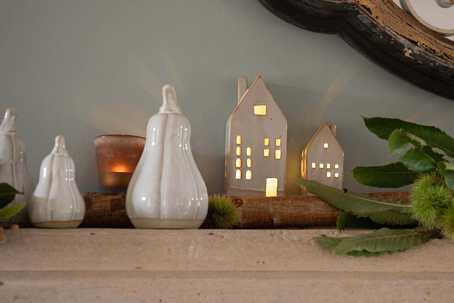 A decorative display likely set in a domestic setting. Resting on a stone base, various objects are arrayed against a wall with a soft gray-green hue. On the left side, three white ceramic figures are seen, shaped like stylized pumpkins or pears with smooth, shiny surfaces. Beside these figures sits a candle holder containing a candle, which radiates a warm, orange-like light through the translucent, brown-tinted glass.

In the center and to the right of the image, there are three ceramic houses with a simplistic design, finished in a matte gray shade. They feature cut-out windows and doors that let light shine through, suggesting that candles or lights are placed inside, giving these houses a cozy glow. It appears that these houses are part of a decorative set meant to represent a sort of miniature village. The entire setting is embellished with natural elements such as a branch and several green leaves, giving the scene a fresh look. The light emanating from the candle and the illuminated houses creates an atmosphere of warmth and conviviality, often associated with homey decorations. The overall image is calming and serene.