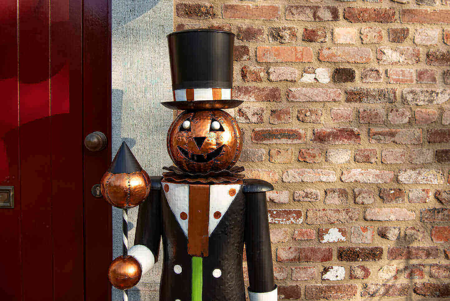 The image displays a metal sculpture of a figure shaped like a pumpkin man set against the background of a brick wall. The pumpkin man is adorned with a top hat and a tuxedo, evoking the style of an old-fashioned gentleman. His face is a carved pumpkin featuring the classic Halloween expression: triangular eyes and a grinning mouth with teeth. The sculpture seems to be made of copper or another metal with a rusty, patinated finish, giving it an antique and handcrafted feel. The figure is dressed in a coat with a white shirt and a brown and white bow tie and holds what appears to be a staff or walking stick in one hand, adorned with spheres of varying sizes. Beside the pumpkin man, a portion of a door painted in a deep red color is visible, providing a striking contrast with the brown of the bricks. The sculpture itself is positioned on a threshold, likely serving as a welcoming or decorative element for guests.