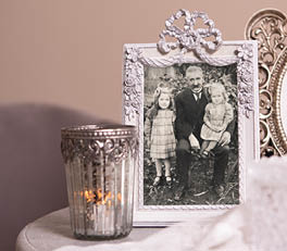 Two photo frames sit on a wooden surface, likely a part of a domestic interior. The left frame boasts a soft blue color with decorative bird and floral details in relief on the sides. Inside, there appears to be a small artwork or printed image featuring yellow flowering plants. The right frame is larger, sporting a classic white finish with intricate, floral-like patterns in relief at the corners and along the edges. It showcases an image or artwork with a delicate floral design, harmonizing with the nature-themed motif of the smaller frame.

Behind the frames, we partially see a woven object, possibly a basket or wall decoration, and on the right side, there is a piece of rope tied in a simple bow. Flanking the frames on both sides are green houseplants in terracotta pots, further enhancing the natural and rustic ambiance of the composition. The soft colors and use of natural materials and motifs suggest an appreciation for nature and a cozy, soothing atmosphere.