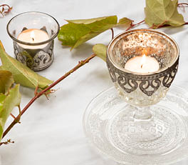 The photo depicts a series of candle holders arranged on a light surface. The candle holders are made of glass and come in various sizes and designs. Two of them are larger and have a coral pink color with a floral relief, giving them a romantic and decorative appearance. The other two are smaller and more neutral in color, one with a transparent pattern and the other with a white and gray design. The soft, natural lighting accentuates the translucent and shimmering nature of the glassware. On the left side, a portion of a green leaf is visible, adding to the natural ambiance of the composition.