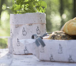 A charming outdoor scene with a rustic vibe unfolds. A wooden bench is nestled against a backdrop of vertical wooden planks, likely a part of a fence or outdoor shed. Resting on the bench is a cushion, inviting with its comfortable appearance, featuring a white background adorned with a subtle olive motif. The cushion boasts a green ruffle edge for an extra touch of color and texture.

On the wooden table beside the bench lies a matching fabric, presumably a napkin or tablecloth, also adorned with the same olive motif. The table itself is set with what appears to be a relaxed outdoor dining or snacking scene, featuring two glasses of wine, a cheese platter with assorted pieces of cheese, and some grapes nearby. Hanging on the right side of the table is a straw hat with a bandana knotted around it, contributing to the rural, laid-back aesthetic. Beneath the table, two wicker baskets further reinforce the pastoral theme. The scene exudes tranquility and comfort, inviting relaxation in the great outdoors.