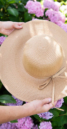 A girl with long, wavy blonde hair, seen from the neck up. She wears a wide-brimmed, beige straw hat adorned with a stylish ribbon featuring feathers and beads. The hat casts a soft shadow over her face, obscuring it partially. She holds a denim shoulder bag with details of small, light flowers on it. The overall vibe exudes a bohemian and summery style, and the girl appears to be posing against a rustic backdrop of white brick, evoking a relaxed and fashionable atmosphere.