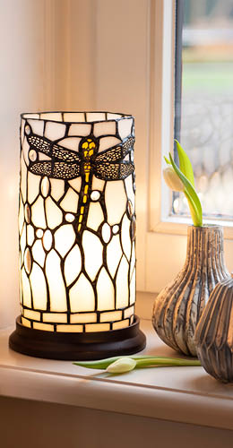 A table lamp reminiscent of one of the Tiffany lamps is depicted. It has a rectangular, tower-like shape with stained glass panels arranged in a geometric manner. The colors of the panels vary from cream in the center to dark blue and black on the sides, creating an elegant color gradient. The lamp is placed on a wooden table or cabinet and features a dark metal frame at the bottom. In the background, another type of Tiffany lamp is visible, with a more traditional, dome-shaped design. The setting exudes a cozy, warm atmosphere with a homely feel, including a wooden cabinet and a plant in the corner of the room.