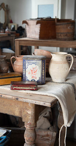 A vintage and rustic composition consisting of various antique objects, displayed on an old, green wooden piece of furniture. At the center sits a black felt hat, resting on a glass tray with a label attached, suggesting it might be for sale in an antique shop or market. Behind the hat is a framed educational poster featuring images and descriptions of various tree species and leaves. To the left of it stands a large glass jar, and partially visible is an antique portrait of a figure, likely of religious nature, framed with an ornate wooden frame. The atmosphere evokes a sense of bygone eras, appealing to history and curiosity enthusiasts.