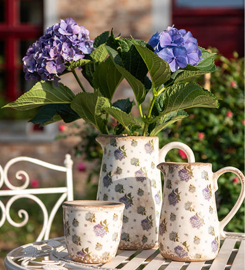 A charming set, consisting of two pitchers and a matching flower pot, all adorned with a delicate floral pattern in shades of lilac and green, imparting a rural feel. The items are displayed on a white garden table with a striped pattern. A full bunch of hydrangeas in various shades of blue adorns the pitcher, adding a vibrant touch of nature. The setting is outdoors, possibly on a porch or in a garden, with the dusky backdrop of a country house and a metal garden bench creating an atmosphere of rustic elegance. It's an idyllic portrayal of outdoor living and relaxation.
