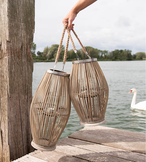 Two lanterns with a natural, wooden appearance, elegantly designed with a narrow top that widens towards the bottom. They feature a rope handle, adding a rustic feel and providing easy portability. The lanterns are being held by a person, of whom only the hand and forearm are visible. This setting is outdoors, close to a body of water, as indicated by the background where a swan peacefully floats, creating a serene and calming effect. It appears to be a perfect accessory for an evening by the water or for creating a cozy atmosphere in any outdoor space.