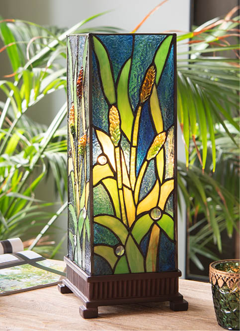 Eine Tischlampe mit einem Bleiglas-Design, die auf einem hölzernen Fuß steht. Die Glaskunstwerke zeigen lebendige Farben und stellen ein Bild von blühenden Pflanzen und Blättern dar, hauptsächlich in Grüntönen, Gelb und Orange, was ein warmes und natürliches Erscheinungsbild ergibt. Die Lampe befindet sich drinnen in einem Raum mit üppigen grünen Pflanzen im Hintergrund, was zur botanischen Atmosphäre der Szene beiträgt. Der Stil der Lampe verleiht dem Inneren einen künstlerischen und antiken Flair, und das durch sie gefilterte Licht würde einen beruhigenden Schein im Raum erzeugen.