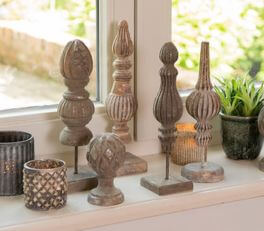 A collection of decorative objects adorns a windowsill. There are a total of seven items, all seemingly crafted from wood with different designs. They are placed on small pedestals, resembling trophies or decorative finials. The colors are predominantly earthy tones, with variations of brown and gray, giving off a rustic and antique feel. Alongside these wooden objects are two candle holders with intricate carvings that allow light to pass through in a charming manner. Behind these objects sits a potted houseplant, adding a touch of green to the scene and providing a refreshing contrast to the predominantly brown hues. The natural light streaming through the window imparts a warm and inviting ambiance to the entire arrangement.