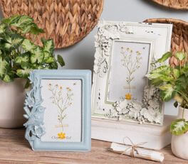 Two photo frames sit on a wooden surface, likely a part of a domestic interior. The left frame boasts a soft blue color with decorative bird and floral details in relief on the sides. Inside, there appears to be a small artwork or printed image featuring yellow flowering plants. The right frame is larger, sporting a classic white finish with intricate, floral-like patterns in relief at the corners and along the edges. It showcases an image or artwork with a delicate floral design, harmonizing with the nature-themed motif of the smaller frame.

Behind the frames, we partially see a woven object, possibly a basket or wall decoration, and on the right side, there is a piece of rope tied in a simple bow. Flanking the frames on both sides are green houseplants in terracotta pots, further enhancing the natural and rustic ambiance of the composition. The soft colors and use of natural materials and motifs suggest an appreciation for nature and a cozy, soothing atmosphere.
