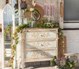 A charming and rustic corner adorned with vintage flair catches the eye. At its heart sits an aged, cream-colored wooden dresser, adorned with gracefully carved details and weathered finishing, lending it an antique allure. The drawers appear exquisitely crafted with floral motifs.

Atop the dresser, a decorative arrangement is meticulously curated with various elements. A collection of candles in varying heights sits elegantly in holders. Amidst the candles rests a small bouquet of flowers in a vase and a round, wooden mirror with rustic charm. Hanging on one side of the dresser is a wooden arch-shaped decoration, reminiscent of a church window, adding a touch of gothic or rural charm.

The dresser is partially adorned with a garland of green leaves and berries, casually draped to introduce a natural, organic element to the setting. On the left side of the floor sits a wooden stool adorned with a bundle of purple flowers, enhancing the rural and romantic style of the scene. It all stands against a wall partially made of bricks and partially painted white wooden panels, accentuating the environment's character. The natural light and choice of decorations craft a warm, inviting, and somewhat nostalgic ambiance.