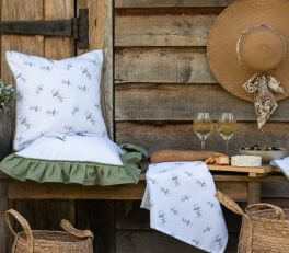 A charming outdoor scene with a rustic vibe unfolds. A wooden bench is nestled against a backdrop of vertical wooden planks, likely a part of a fence or outdoor shed. Resting on the bench is a cushion, inviting with its comfortable appearance, featuring a white background adorned with a subtle olive motif. The cushion boasts a green ruffle edge for an extra touch of color and texture.

On the wooden table beside the bench lies a matching fabric, presumably a napkin or tablecloth, also adorned with the same olive motif. The table itself is set with what appears to be a relaxed outdoor dining or snacking scene, featuring two glasses of wine, a cheese platter with assorted pieces of cheese, and some grapes nearby. Hanging on the right side of the table is a straw hat with a bandana knotted around it, contributing to the rural, laid-back aesthetic. Beneath the table, two wicker baskets further reinforce the pastoral theme. The scene exudes tranquility and comfort, inviting relaxation in the great outdoors.