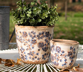 An idyllic and sunny setting with two flower pots on a wooden surface, in front of a window with a weathered appearance. The pots have a vintage design with a cream base and a flower pattern featuring roses in pink and green hues. The light shining through the window gives the scene a warm and inviting atmosphere. The setting exudes rustic charm, and it appears to be a part of a rural interior.