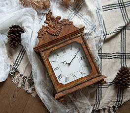 The photo depicts an elegant mantel clock with a vintage appearance. The clock features a classic face against a white background. The clock face is surrounded by an elaborately decorated frame with swirling, baroque motifs. The frame has a weathered, white color that adds to its antique character. The clock is placed on what appears to be a windowsill, with sunlight streaming through a window with a weathered frame. This gives the scene a warm, inviting atmosphere and accentuates the texture of the clock's metal frame.