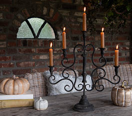 The photo depicts a series of candle holders arranged on a light surface. The candle holders are made of glass and come in various sizes and designs. Two of them are larger and have a coral pink color with a floral relief, giving them a romantic and decorative appearance. The other two are smaller and more neutral in color, one with a transparent pattern and the other with a white and gray design. The soft, natural lighting accentuates the translucent and shimmering nature of the glassware. On the left side, a portion of a green leaf is visible, adding to the natural ambiance of the composition.