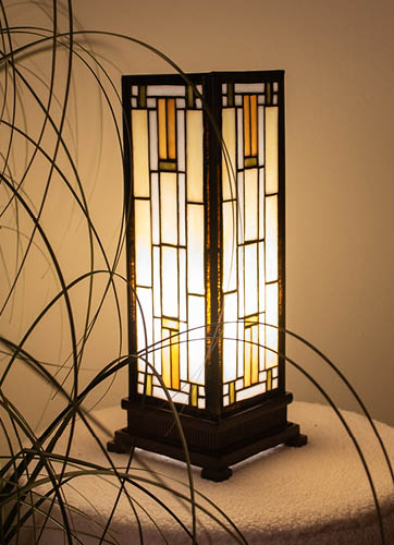 A table lamp reminiscent of one of the Tiffany lamps is depicted. It has a rectangular, tower-like shape with stained glass panels arranged in a geometric manner. The colors of the panels vary from cream in the center to dark blue and black on the sides, creating an elegant color gradient. The lamp is placed on a wooden table or cabinet and features a dark metal frame at the bottom. In the background, another type of Tiffany lamp is visible, with a more traditional, dome-shaped design. The setting exudes a cozy, warm atmosphere with a homely feel, including a wooden cabinet and a plant in the corner of the room.