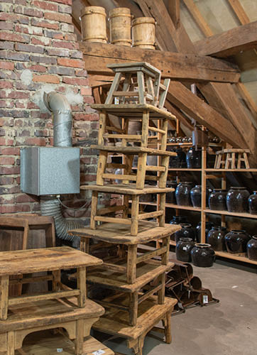 Une composition vintage et rustique, composée de différents objets d'antiquités, disposés sur un vieux meuble en bois vert. Au centre se trouve un chapeau en feutre noir, posé sur un plateau en verre avec une étiquette, suggérant qu'il pourrait être à vendre dans une brocante ou sur un marché. Derrière le chapeau se trouve une affiche éducative encadrée avec des images et des descriptions de différentes espèces d'arbres et de feuilles. À gauche se trouve un grand tonneau en verre et une partie d'un portrait ancien d'une figure, probablement de nature religieuse, encadré avec un cadre en bois décoré. L'atmosphère est celle des temps révolus, et parle aux amateurs d'histoire et de curiosités.