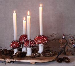 The photo depicts a series of candle holders arranged on a light surface. The candle holders are made of glass and come in various sizes and designs. Two of them are larger and have a coral pink color with a floral relief, giving them a romantic and decorative appearance. The other two are smaller and more neutral in color, one with a transparent pattern and the other with a white and gray design. The soft, natural lighting accentuates the translucent and shimmering nature of the glassware. On the left side, a portion of a green leaf is visible, adding to the natural ambiance of the composition.