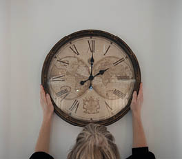 The photo depicts an elegant mantel clock with a vintage appearance. The clock features a classic face against a white background. The clock face is surrounded by an elaborately decorated frame with swirling, baroque motifs. The frame has a weathered, white color that adds to its antique character. The clock is placed on what appears to be a windowsill, with sunlight streaming through a window with a weathered frame. This gives the scene a warm, inviting atmosphere and accentuates the texture of the clock's metal frame.