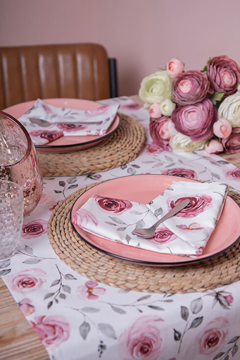 Twee ronde placemats, twee roze borden, een wit tafelkleed met bloemen en drinkglazen