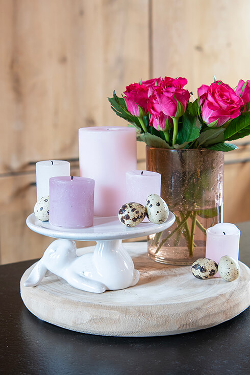 Un piatto a forma di coniglio bianco con candele dai colori pastello e un vaso di rose su un vassoio di legno
