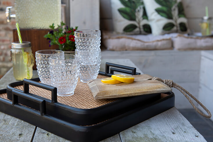 Un vassoio con finitura in vimini e bordi neri, con vari bicchieri e una tavola da taglio con limoni affettati