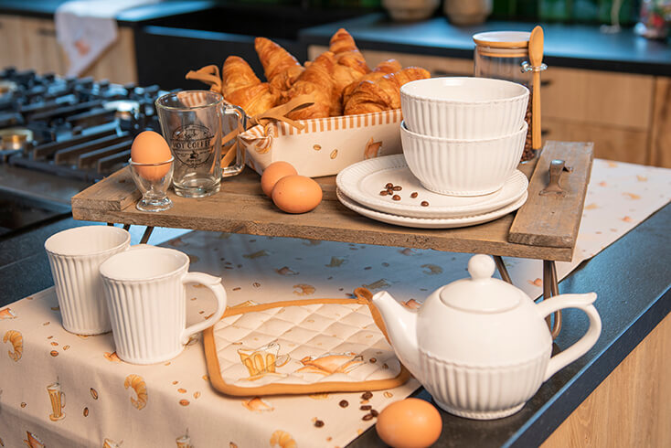 Een houten dienblad met ontbijt en servies, waaronder een broodmandje, kommetjes, theepotten, mokken en een glazen eierdopje