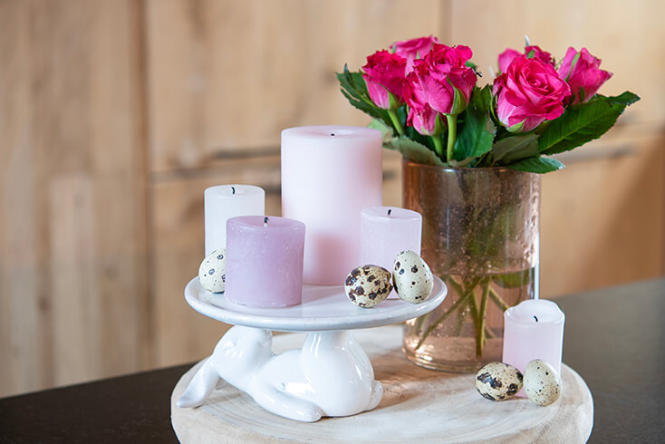 Een konijnenschaal met pastelkleurige kaarsen en een roze vaas met rozen op een houten dienblad met enkele kwarteleieren