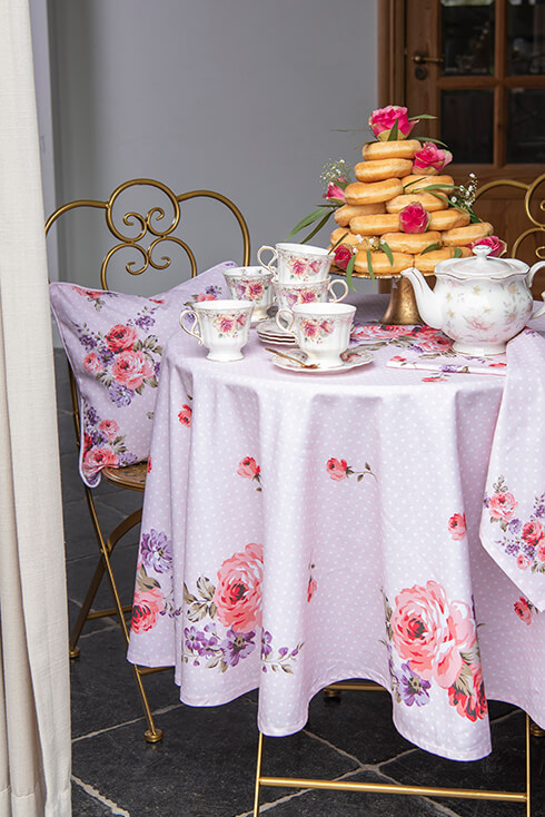 Een gedekt bistrotafeltje met een roze ronde tafelkleed, sierkussen en romantisch servies, waaronder kop en schotels en een theepot en een gouden taartplateau met gestapelde donuts