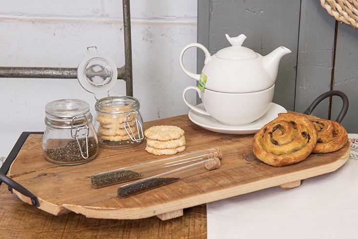 Ein Holztablett mit einem weißen Tea-for-One-Set, zwei Glasvorratsdosen und Keksen