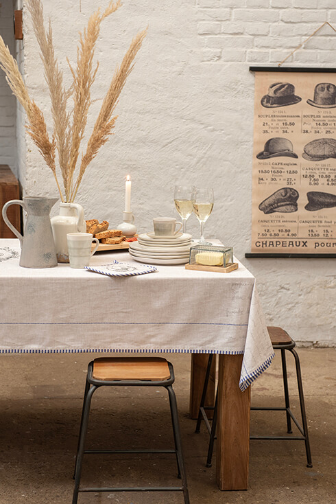 Une table dressée à la campagne avec une nappe en lin, des tabourets, de la vaisselle, un beurrier, des pichets, des fleurs des champs et des verres à vin