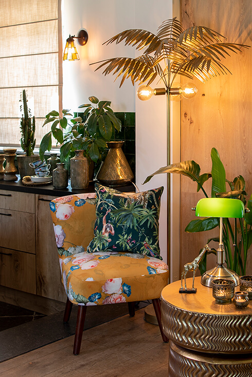 Ein botanisches Interieur mit einem Sessel mit Blumenmuster, einer Stehlampe in Form eines Palmenbaums, einer Bankerlampe, einem dekorativen Kissen, einer Giraffenfigur und einem goldfarbenen Beistelltisch