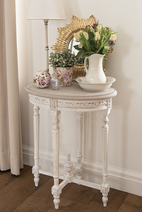 Un tavolino rustico con sopra un specchio da parete dorato, una lampada da tavolo shabby chic, un catino bianco, un vaso per fiori, una sfera decorativa e un vasetto di vetro