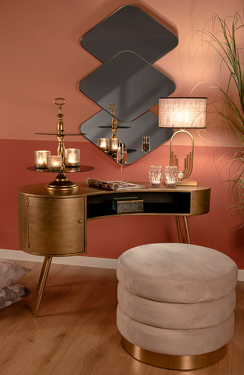 Una scrivania dorata con un poggiapiedi, e sulla scrivania c'è una lampada da tavolo, una mensola e un calice di champagne, e al muro c'è uno specchio da parete moderno