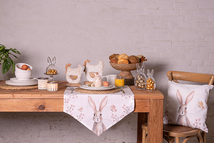 Une table de Pâques dressée avec un chemin de table, de la vaisselle, un plateau à gâteaux, des statues de poules et des bocaux de stockage