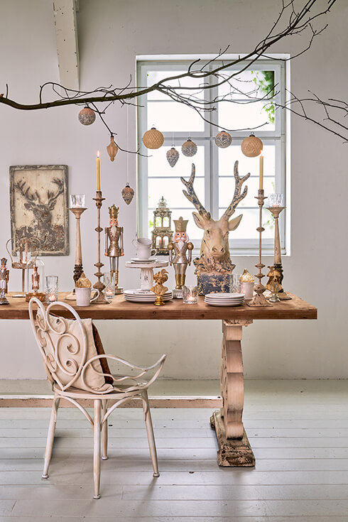 Une table de salle à manger shabby chic avec des décorations de Noël, de la vaisselle, des statues et une chaise en fer romantique avec un coussin décoratif