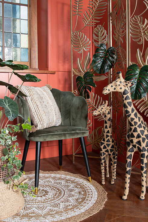 Une chaise verte avec un coussin décoratif à côté de deux figurines de girafe, et derrière eux, un séparateur de pièce doré avec un tapis tissé et de la verdure artificielle
