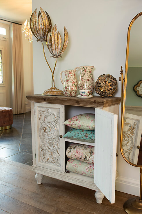 Un mobile da cucina rustico bianco con cuscini decorativi e copriletti sopra, e su di esso si trova una lampada da tavolo, brocche decorative, tazze e piattini e una sfera decorativa