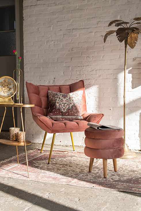 Una poltrona rosa con un poggiapiedi rosa, un grande tappeto e un tavolino da salotto dorato con una lampada da terra