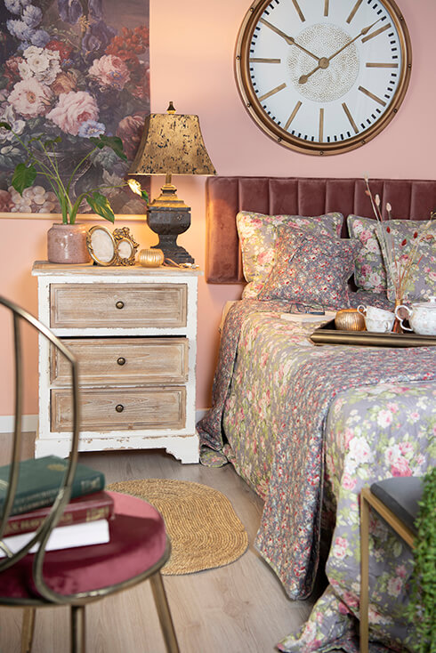 Un letto rifatto con copriletti e cuscini, e accanto si trova una cassettiera di legno con una lampada da tavolo, un vaso per fiori e cornici per foto
