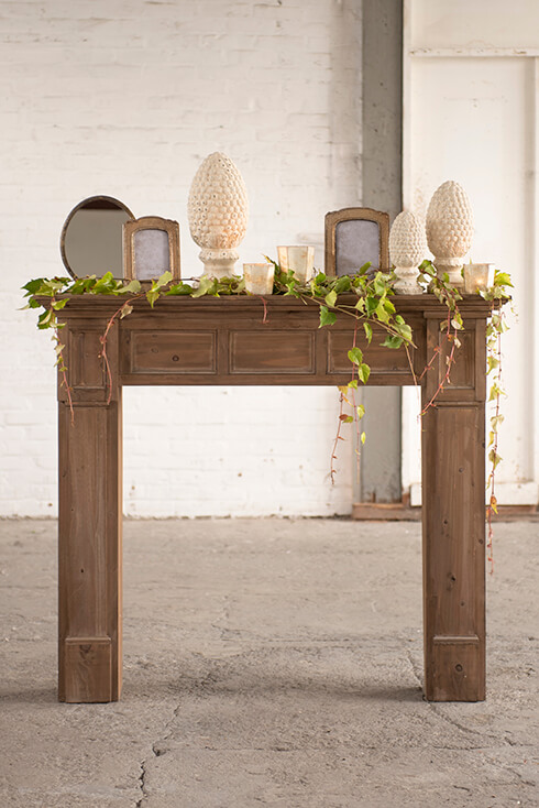 Ein brauner Kaminsims mit drei dekorativen Glöckchen, Bilderrahmen, Teelichthaltern und einem Tischspiegel