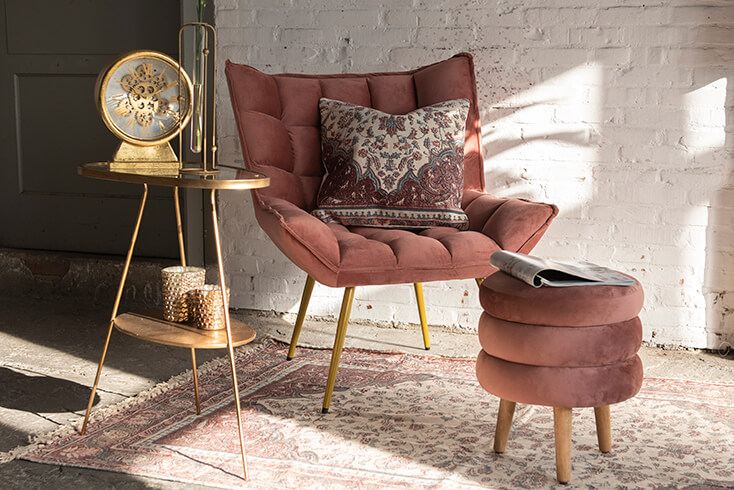 Una poltrona rosa con un cuscino decorativo e un poggiapiedi rosa con un tavolino dorato su cui si trova un orologio dorato, un vaso e portatealight, con un tappeto sul pavimento